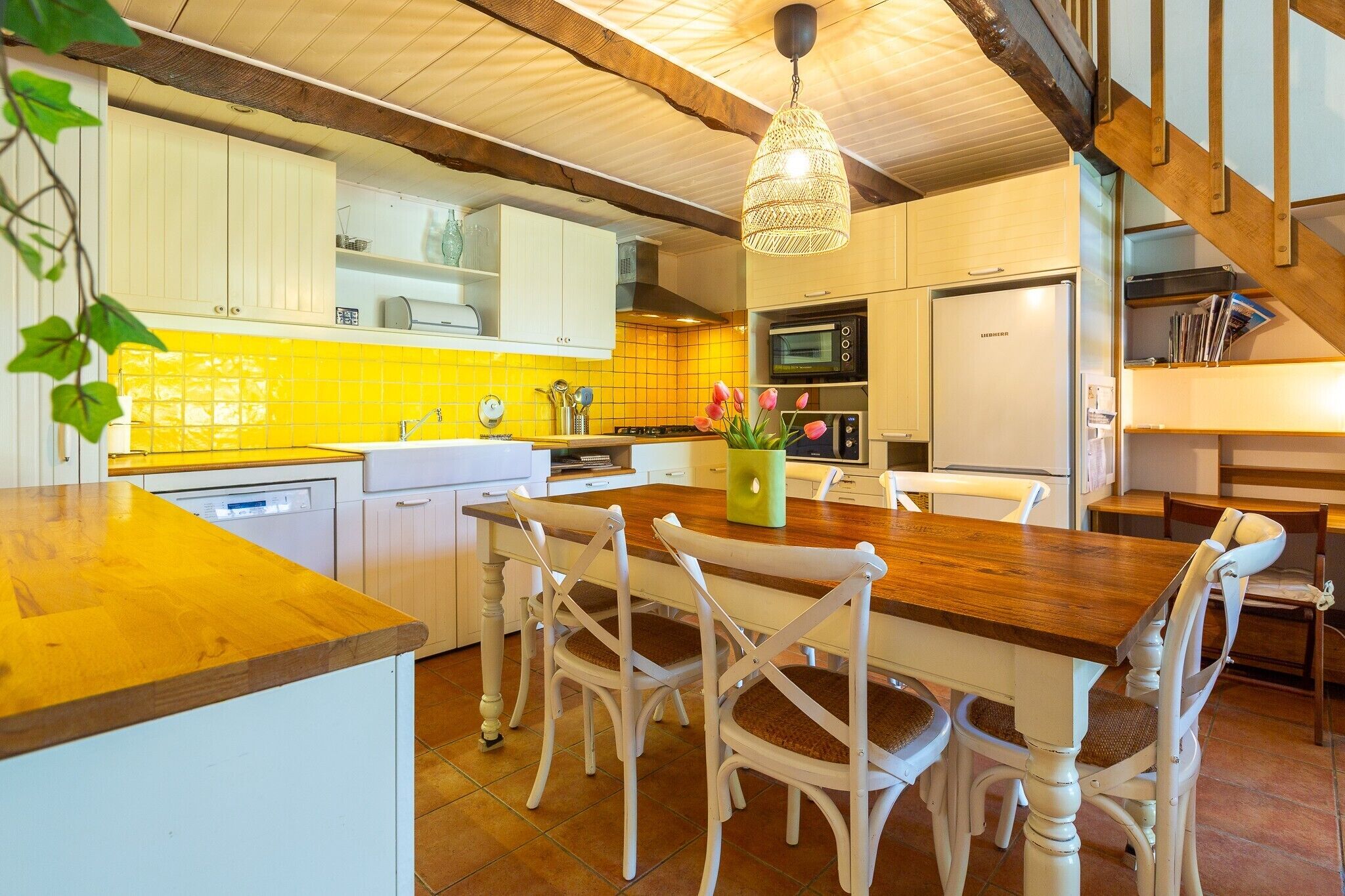 A kitchen with a wooden table and white chairs, a hanging light fixture, and a yellow-tiled backsplash.