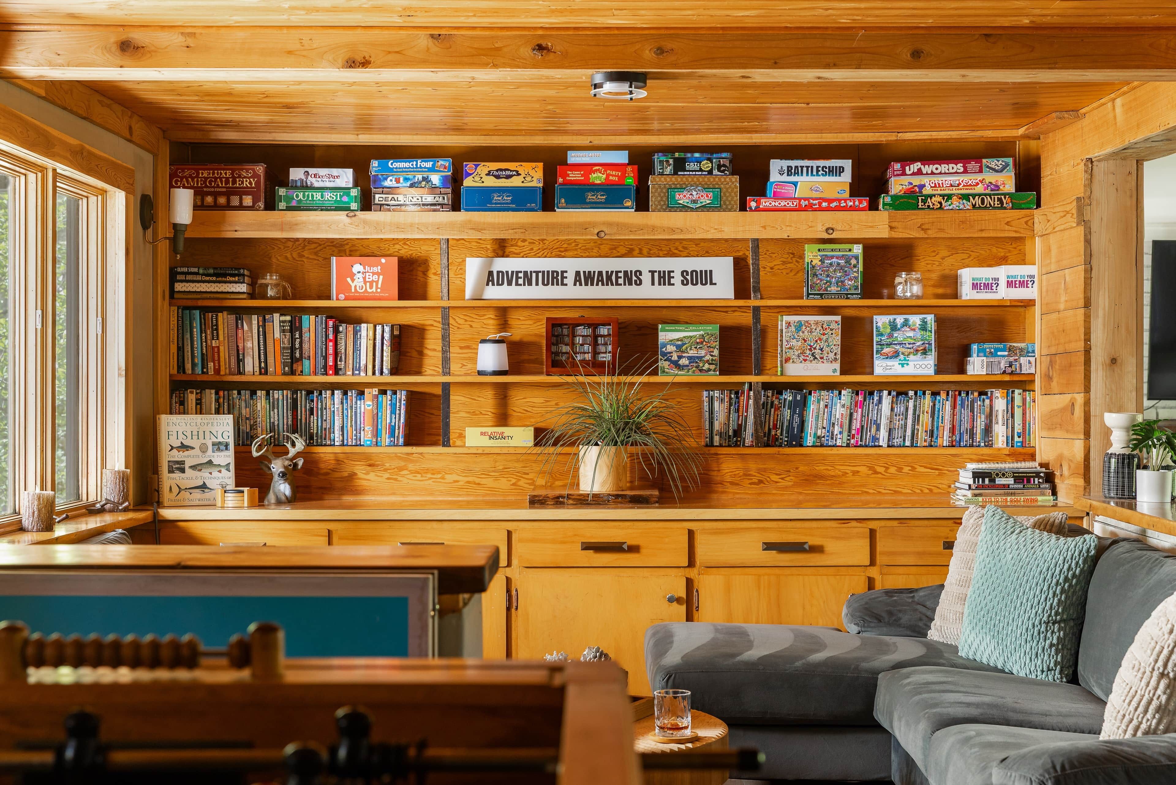 A cozy room with wooden shelves filled with board games, a bookshelf with various books, a comfortable gray sofa, and a wooden desk with a plant.