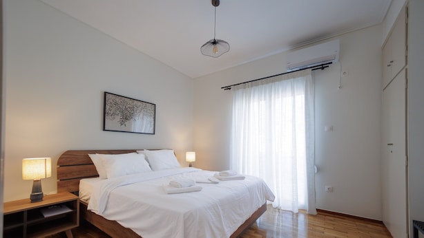 Een slaapkamer met een houten bed, witte beddengoed, een nachtkastje met een lamp, een plafondlamp en een raam met gordijnen.
