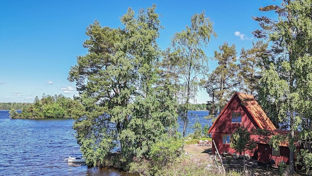 Une cabane au bord d’un lac, avec un toit rouge, entourée d’arbres et sous un ciel bleu clair.