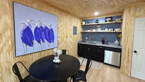 Un coin repas avec une table ronde et des chaises, une cuisine avec des meubles en bois et un dosseret carrelé, et un grand tableau représentant des oiseaux bleus sur le mur.