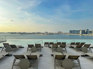 A poolside area with lounge chairs and tables, overlooking a body of water and a distant cityscape. - Beachfront Bliss 2Bedroom in Grand Bleu (Emaar Beachfront)