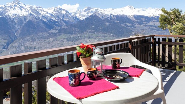 Un balcón con una mesa dispuesta para dos personas, con vistas a un paisaje montañoso.