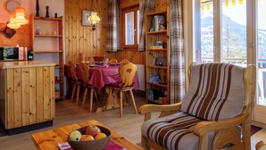 Un interior de cabaña acogedor con comedor, una silla de madera, una mesa con un tazón de fruta y vistas a las montañas a través de amplias ventanas.