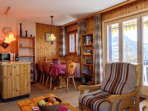 A cozy cabin interior with a dining area, a wooden chair, a table with a bowl of fruit, and a view of mountains through large windows. - Edelweiss by Interhome (Veysonnaz)