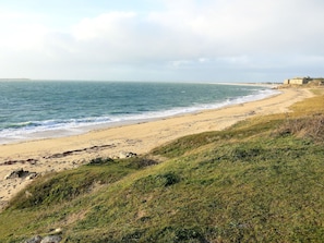 A coastal landscape with a sandy beach, grassy terrain, and a distant building.