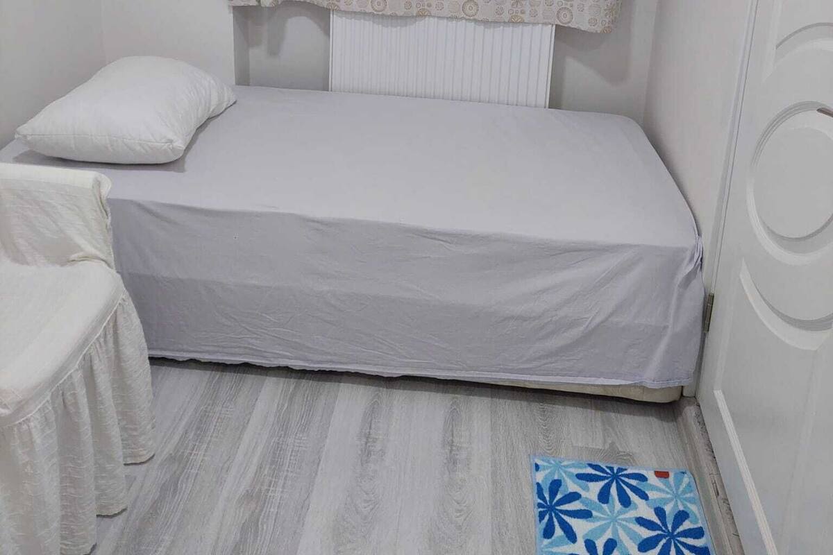 Una habitación pequeña con una cama individual, una cortina estampada, un almohadón blanco y un tapete floral azul.