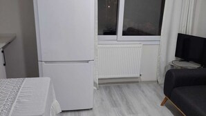 A room with a white refrigerator, a table with a tablecloth, a bed, a window with curtains, and a radiator. - Alaçatı Evi (Cesme)