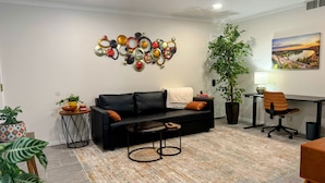 Un salon moderne avec un canapé en cuir noir, une table basse, une plante en pot, une œuvre d’art murale et un bureau avec une chaise.