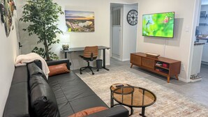 Un salon moderne avec un canapé en cuir noir, une table basse en verre, un meuble TV en bois, un bureau avec une chaise et une grande fenêtre ornée d’un tableau.