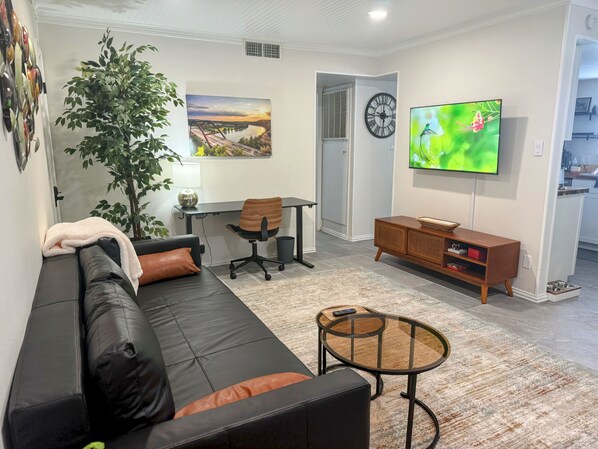 A modern living room with a black leather sofa, a glass coffee table, a wooden TV stand, a desk with a chair, and a large window with a painting. - Remodeled SoCo Home Near Downtown (Austin)