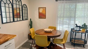 Un coin repas avec une table ronde, des chaises jaunes, un buffet en bois, un tapis en peau de vache, un cactus et une fenêtre avec des stores.