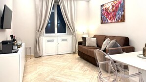A modern living room with a brown sofa, a dining table with chairs, a TV mounted on the wall, and a window with curtains. - Ninfa Maison Rome | Piazza Navona - Coronari (Roma)
