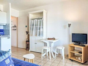 A compact living space with a kitchenette, a dining area with a table and chairs, a TV on a wooden stand, and a wardrobe with a curtain. - Le Grand Large by Interhome (Six Fours La Coudoulière)