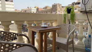 A balcony with wicker chairs, a small wooden table with a cup, and a view of city buildings. - La Bella Casa hostel (Cairo)