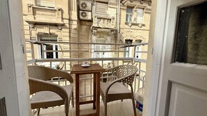 Un balcón con sillas de mimbre, una mesita y una ventana con vistas a un edificio con varias unidades de aire acondicionado.