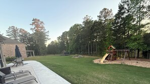 A backyard with a concrete patio, a play area with a slide, and a brick house. - World Cup Ready Spacious 5BR Home Near Atlanta Stadium & Downtown Restaurants (Fairburn)