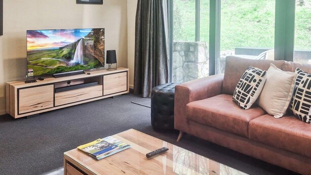 Ein Wohnzimmer mit Fernseher auf Holzfuß, einer Couch mit gemusterten Kissen, einer Fernbedienung auf einem Couchtisch und einer durch ein Fenster sichtbaren Steinwand.