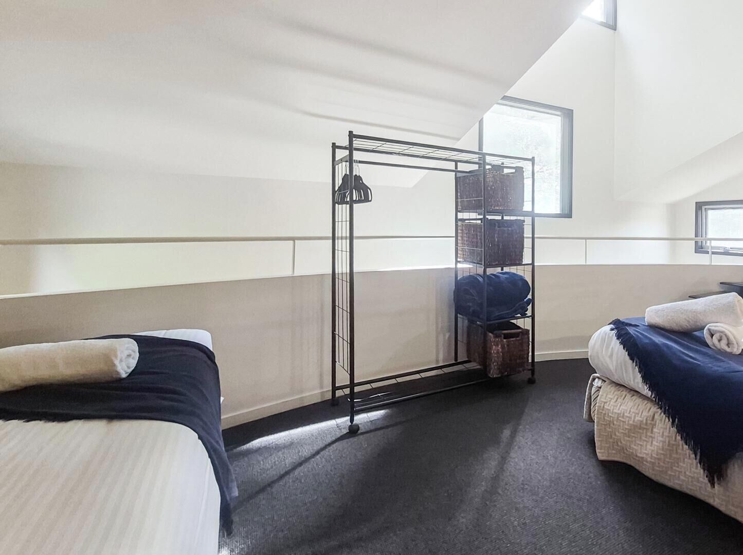 Une chambre d’hôtel avec deux lits, un étagère métallique avec des paniers, une fenêtre et un luminaire au plafond.