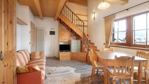 Ein Holzinterieur mit Treppe, einer Couch, einem Esstisch und Blick durch ein Fenster.