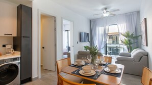 A modern kitchen with a built-in washing machine, a dining area with a table set for four, and a living area with a sofa and a plant. - Pool View, Gym, and Artificial Beach (Punta Cana)