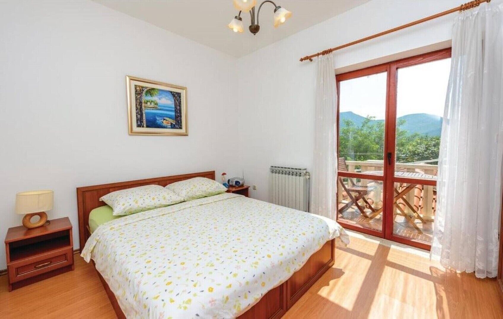 A bedroom with a bed, a nightstand, a painting, a window with curtains, and a view of greenery.