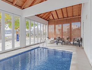 Une piscine intérieure avec des parois en verre transparent, un plafond en bois et un espace extérieur aménagé pour se détendre.
