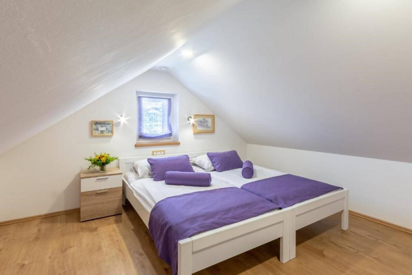 A bedroom with a sloped ceiling, a bed with purple bedding, a nightstand with a vase of flowers, and a window with curtains.