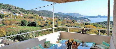 A terrace with a glass table, cushions, and bottles, overlooking a scenic landscape with hills and a body of water.