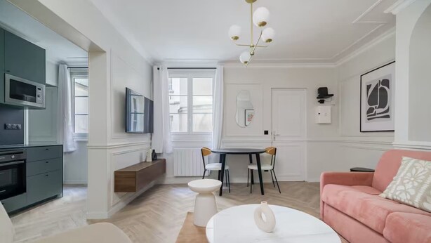 En moderne stue med sofa, sofabord, spisebord med stole, et køkkenområde med mikroovn, og et TV ophængt på væggen.