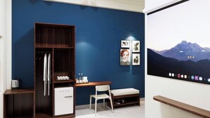 Habitación compacta con pared azul oscuro, armario de madera, escritorio blanco pequeño, una silla, televisor de pantalla plana y un cuadro con paisaje de montaña.