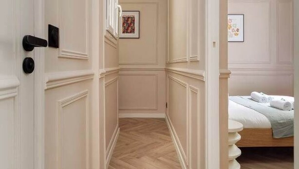 Un couloir aux murs recouverts de lambris blancs et doté d’un plancher en bois, qui mène à une chambre où se trouvent un lit et un luminaire suspendu.