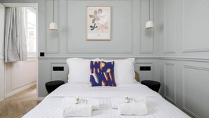 Ein Hotelzimmer mit einem Bett, zwei Nachttischen, einem Fenster mit Vorhängen, einem gerahmten Kunstwerk an der Wand und einem gelben Akzent auf der Bettdecke.