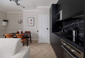 A modern kitchen with dark cabinetry, a microwave, and a stove. There is a dining area with a table and chairs. - PoeticStay - Louvre - Suite Rimbaud (Paris)