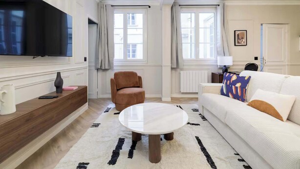 Ein modernes Wohnzimmer mit einem Flachbildfernseher, einem runden Couchtisch, einer Couch mit gemusterten Kissen, einem braunen Sessel und einem großen Fenster.