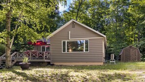 A cabin with a large window, a deck with a red umbrella, and a shed in a wooded area. - Have Lazy Days at Lazy Bear Landing on Glen Lake (Maple City)