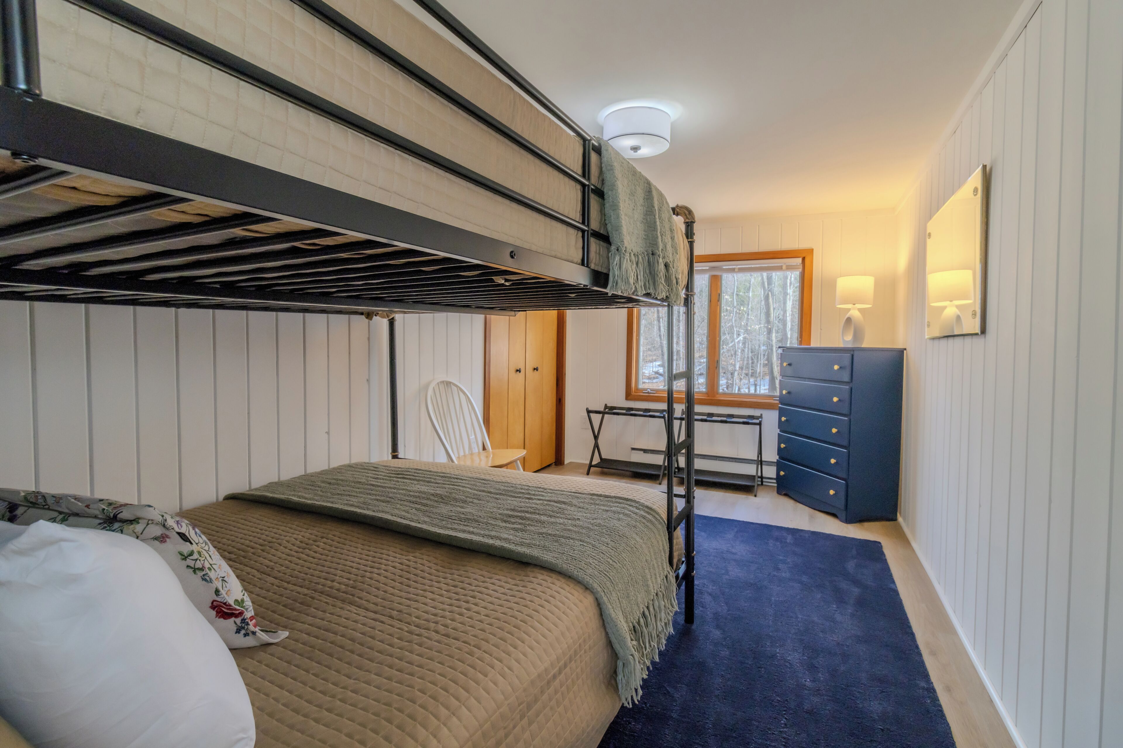 A bunk bed room with a desk, chair, dresser, and window.
