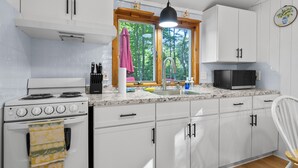 A kitchen with white cabinets, a stove, a microwave, and a window with a view of trees. - Have Lazy Days at Lazy Bear Landing on Glen Lake (Maple City)