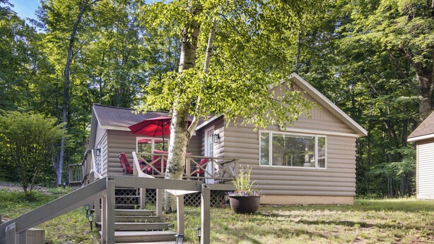 A log cabin with a wooden deck, a red umbrella, and a staircase leading to the entrance. - Have Lazy Days at Lazy Bear Landing on Glen Lake (Maple City)