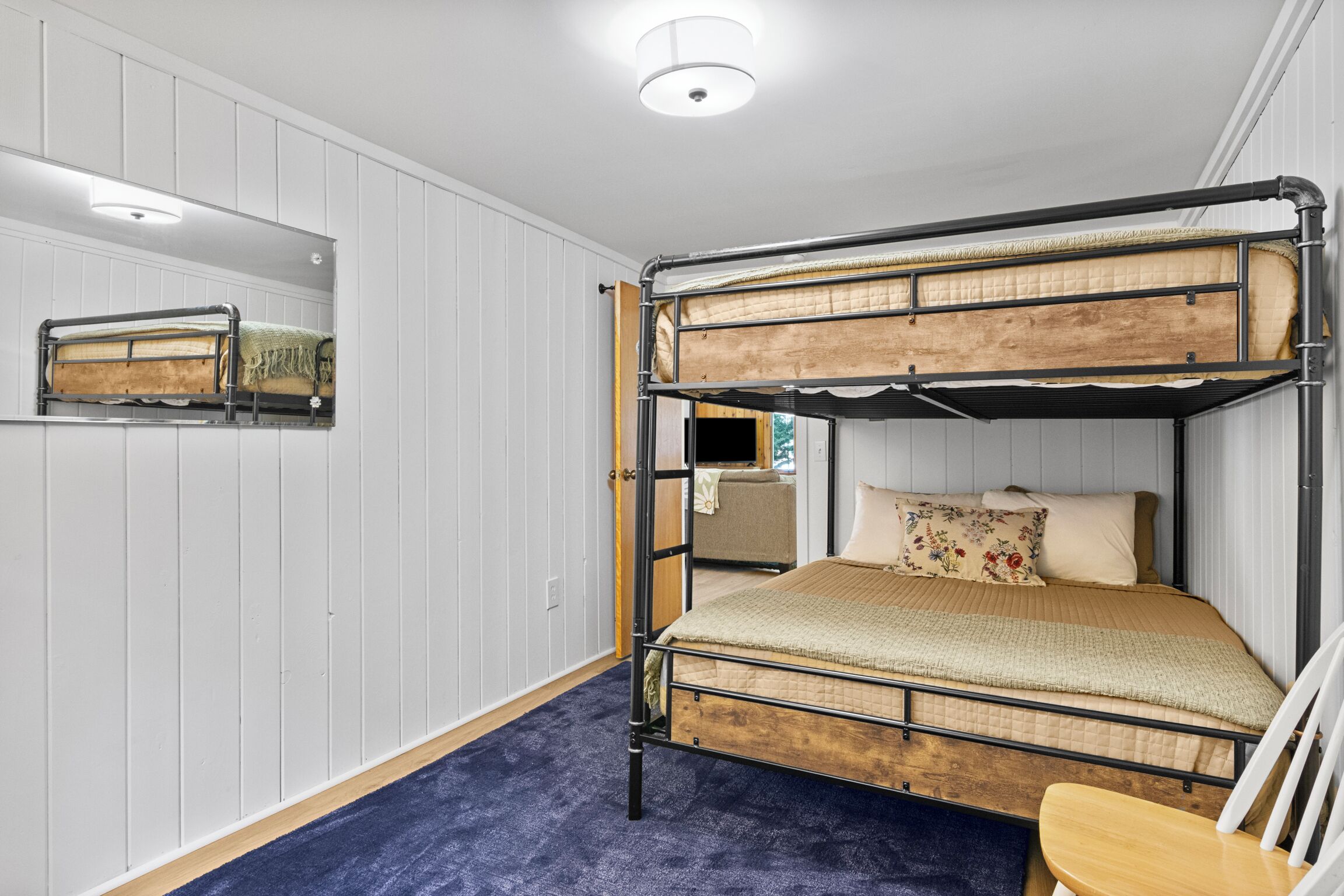 Dormitorio con literas, paredes de paneles blancos, alfombra azul y una silla de madera.