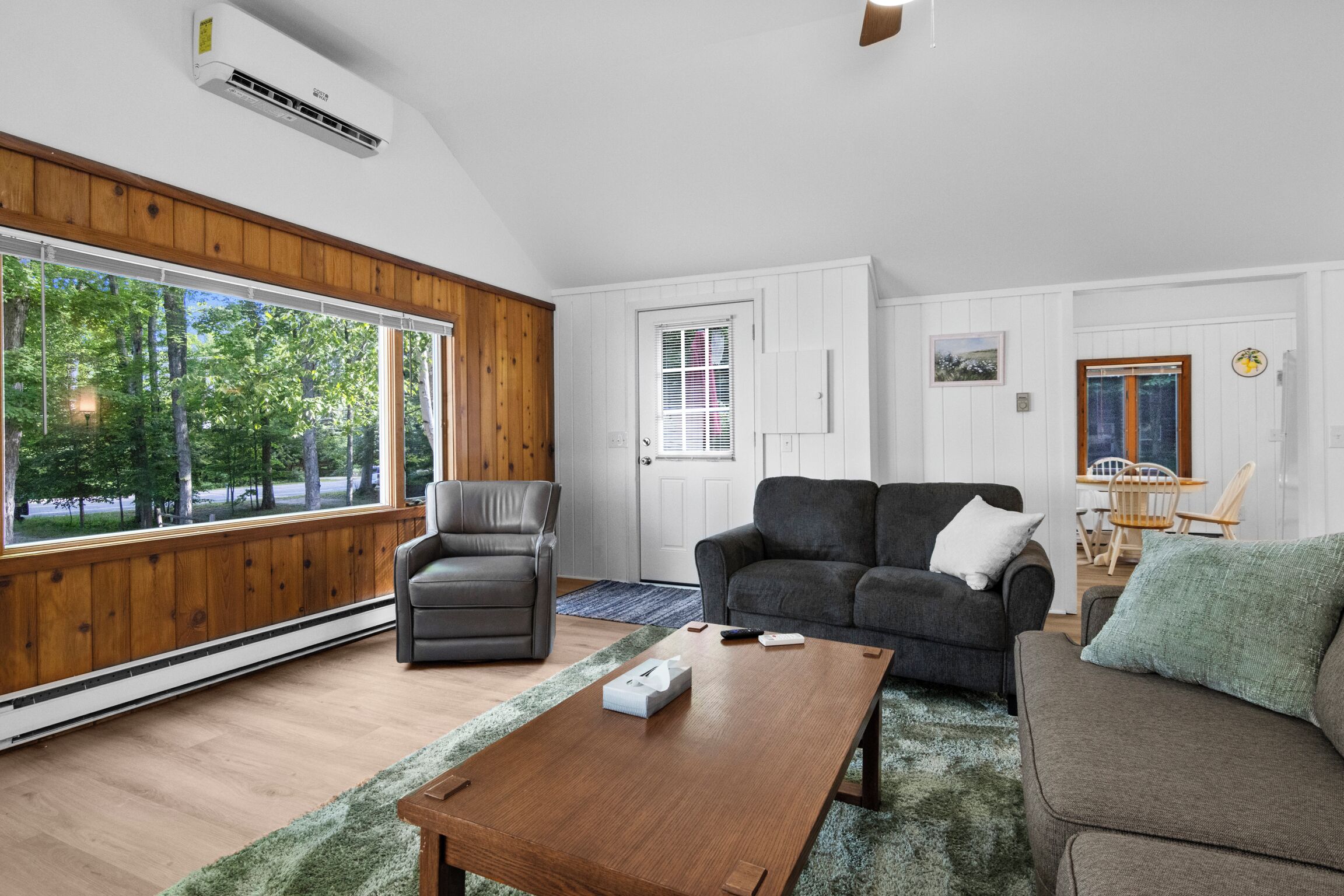Ein Wohnzimmer mit Holzboden, Ledersessel, dunklem Sofa, Couchtisch aus Holz und großen Fenstern mit Blick auf Bäume.