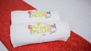 Two folded towels with 'Machu Picchu Land' branding on a bed with a red and white bedspread. - Machu Picchu Land (Aguas Calientes)