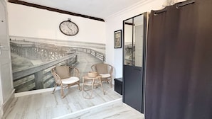 A room with a wooden floor, a black door, a white wall with a clock, and a large window with a view of a bridge.