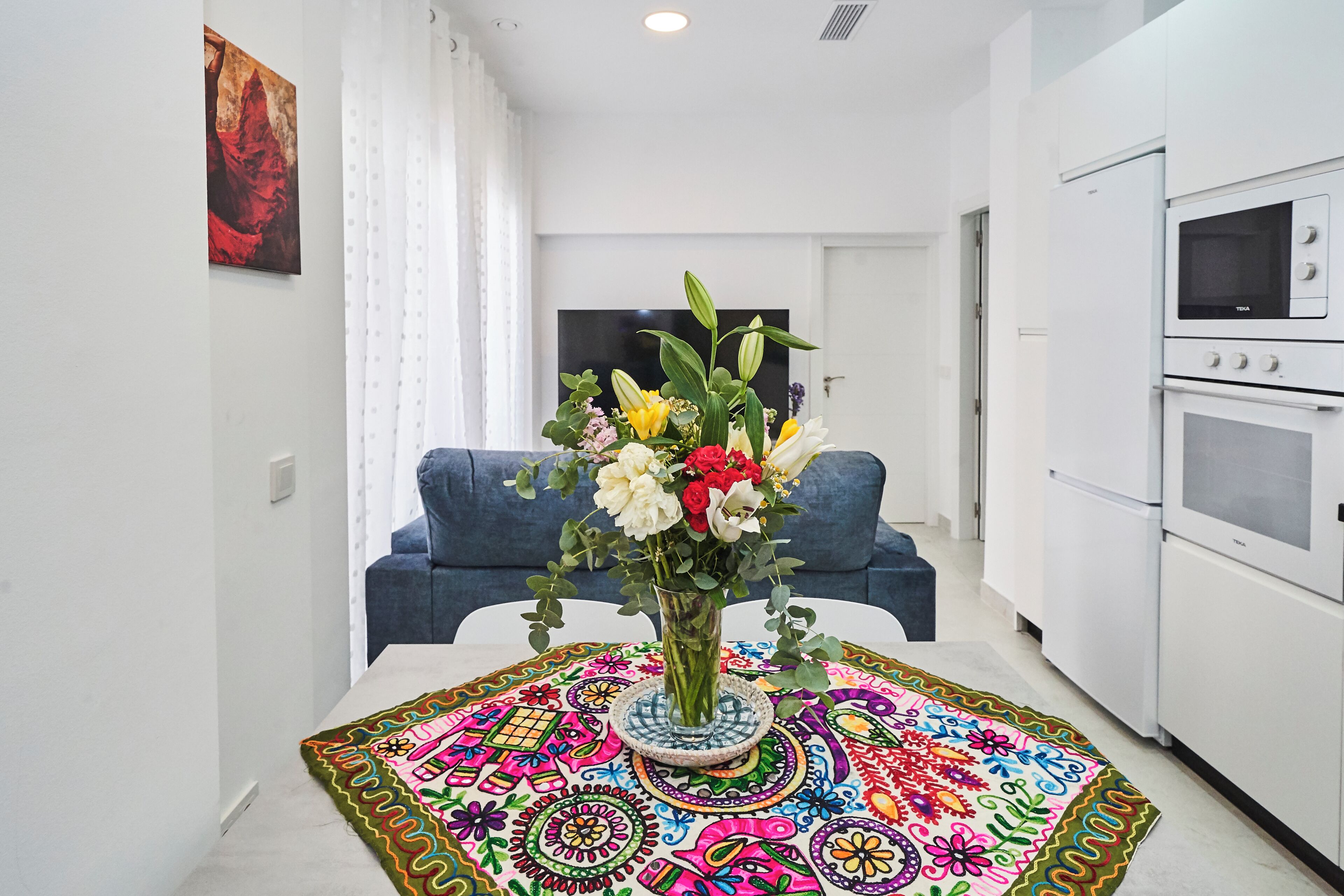 A modern kitchen with white appliances, a dining area with a colorful rug, and a blue sofa in the background.