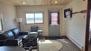 A room with a wooden ceiling, a sofa, a dining table with chairs, a lamp, and a view through a window.