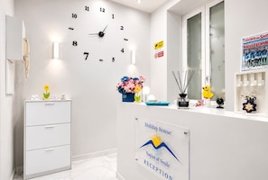 Ein Empfangsbereich mit einer Uhr, einer Blumenvase, einem Schreibtisch mit Telefon und einem Schild mit der Aufschrift 'Holiday house Naples at Smile Reception'.