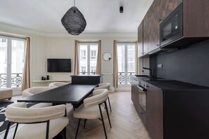 A modern kitchen with a black countertop, a built-in microwave, and a dining area with a black table and white chairs. - Spacious 6PAX flat in the city center (Paris)