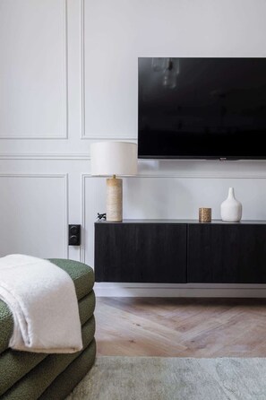 Ein modernes Wohnzimmer mit einem Flachbildfernseher, einer dunklen Holzkommode, einem grünen Sofa und einer weißen Tagesdecke.