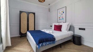 Una camera da letto moderna con un letto, una coperta azzurra, un cuscino rosso, un quadro incorniciato, un cestino nero e una luce a soffitto bianca.