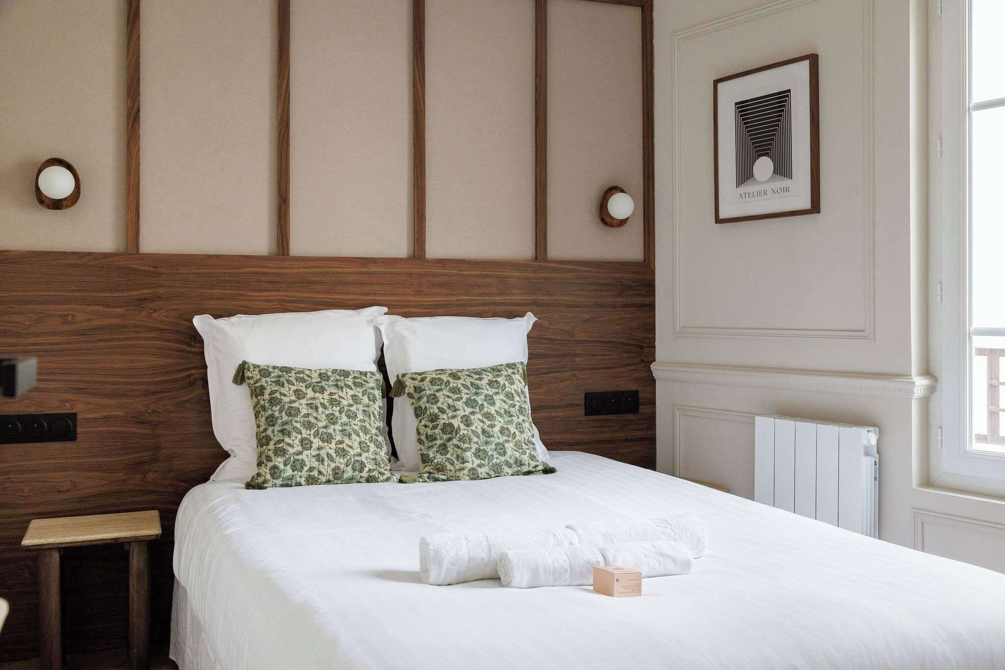 A neatly made bed with white linens and two patterned pillows, a wooden headboard, a framed picture on the wall, and a small bedside table.